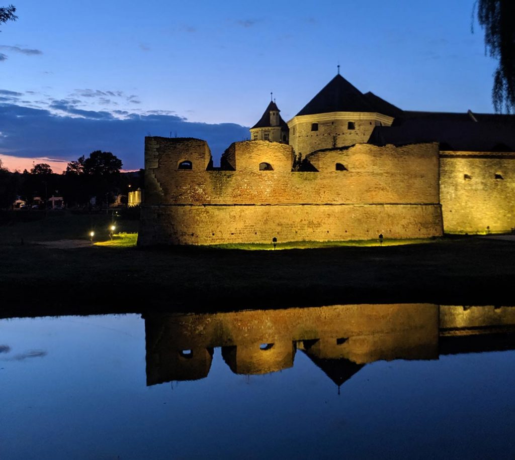 In this photo, a brightly illuminated castle is reflected in the still waters of the moat, set against a backdrop of a late sunset sky.