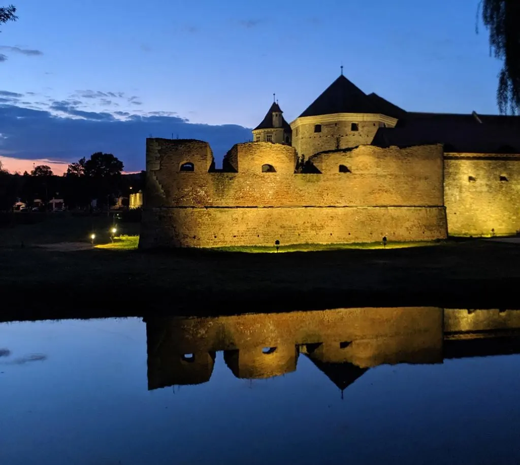 In this photo, a brightly illuminated castle is reflected in the still waters of the moat, set against a backdrop of a late sunset sky.