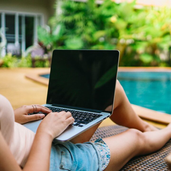 Woman working on a laptop beside a blue green pool, where some digital nomads work.