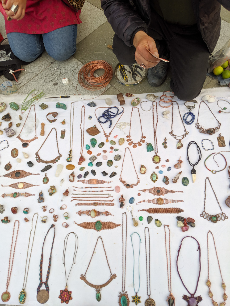 Handmade necklaces spread on a blanket, near a traveler with jewelry making supplies.