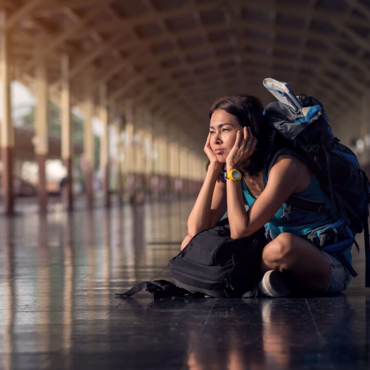 A woman with a weary expression sits on the floor of a train station with her luggage