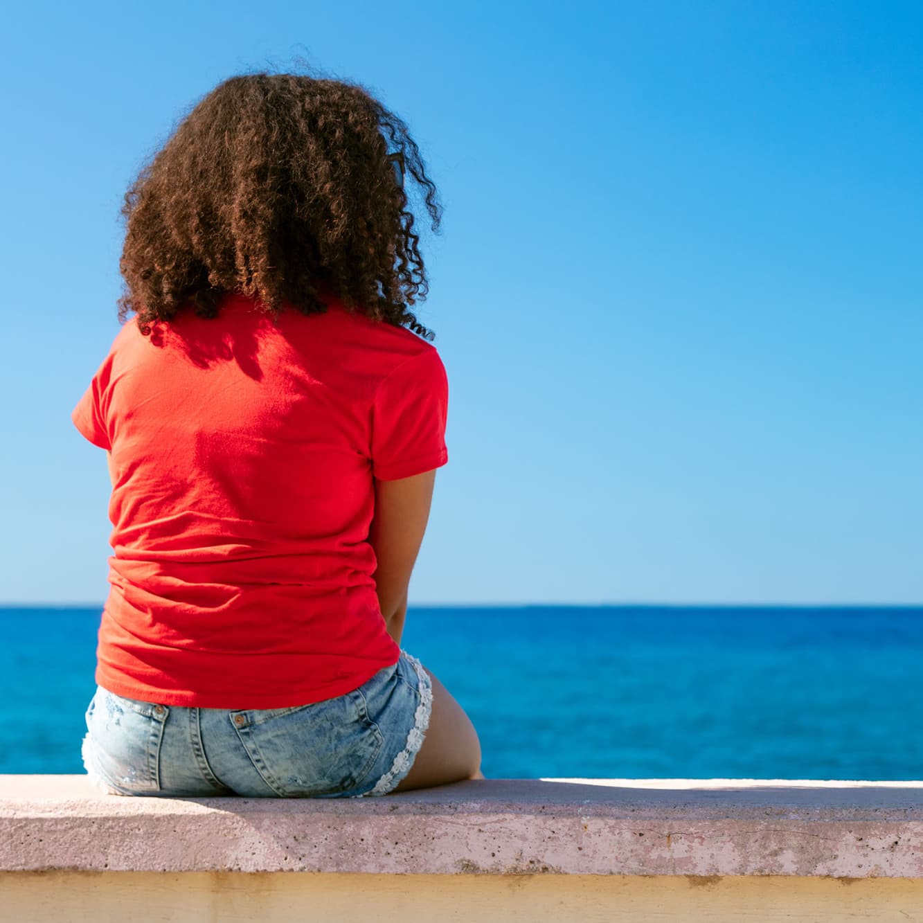 A woman sits on a wall alone near the sea.