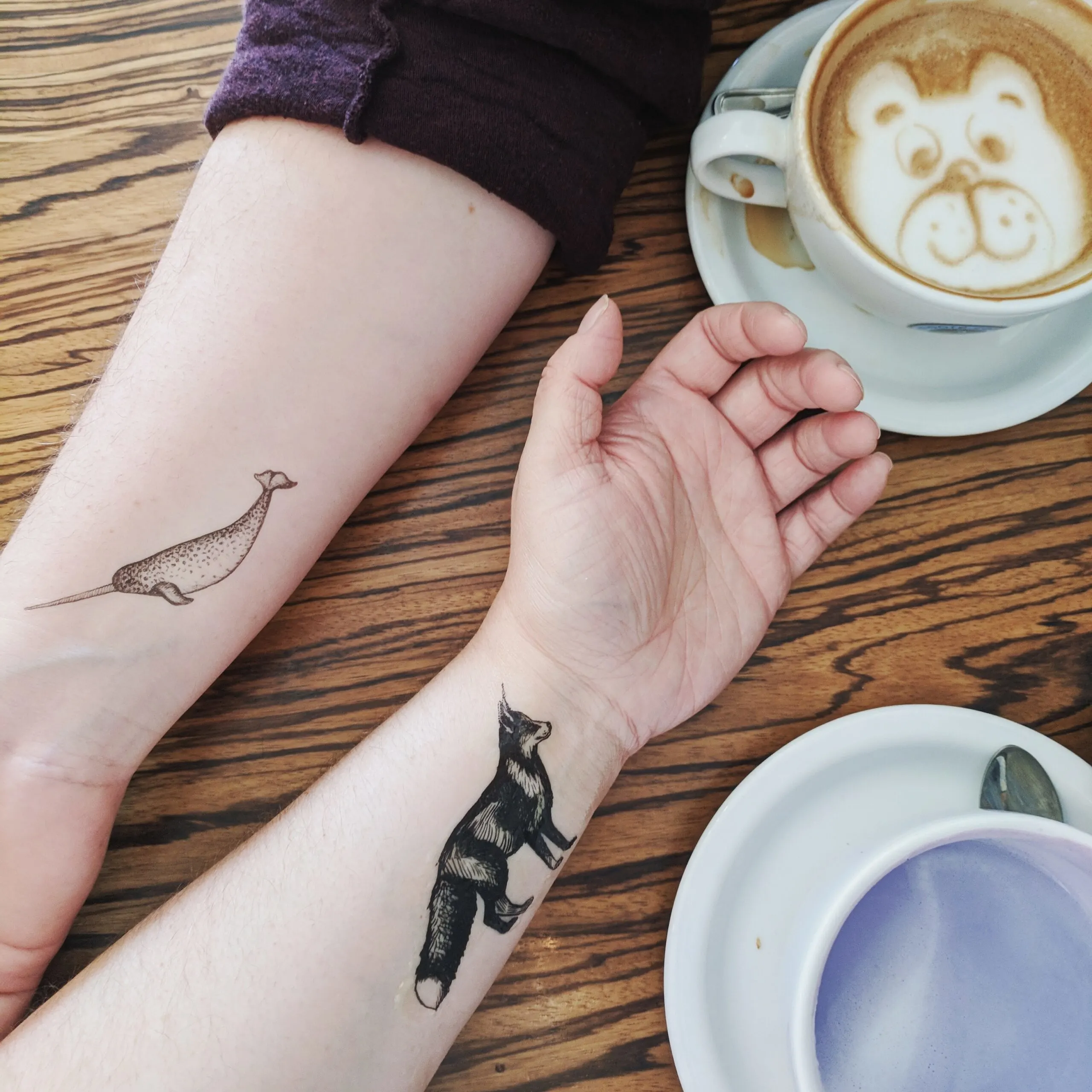 Two tattooed forearms on a table with coffee drinks with latte art.