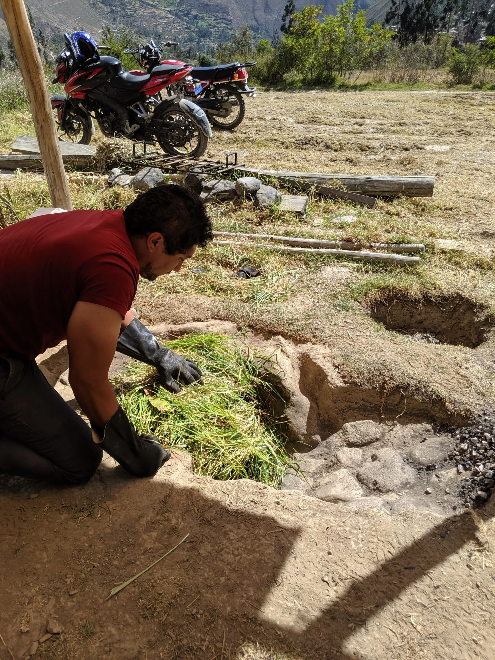 A man spreading grass over the top of an underground oven to create steam.