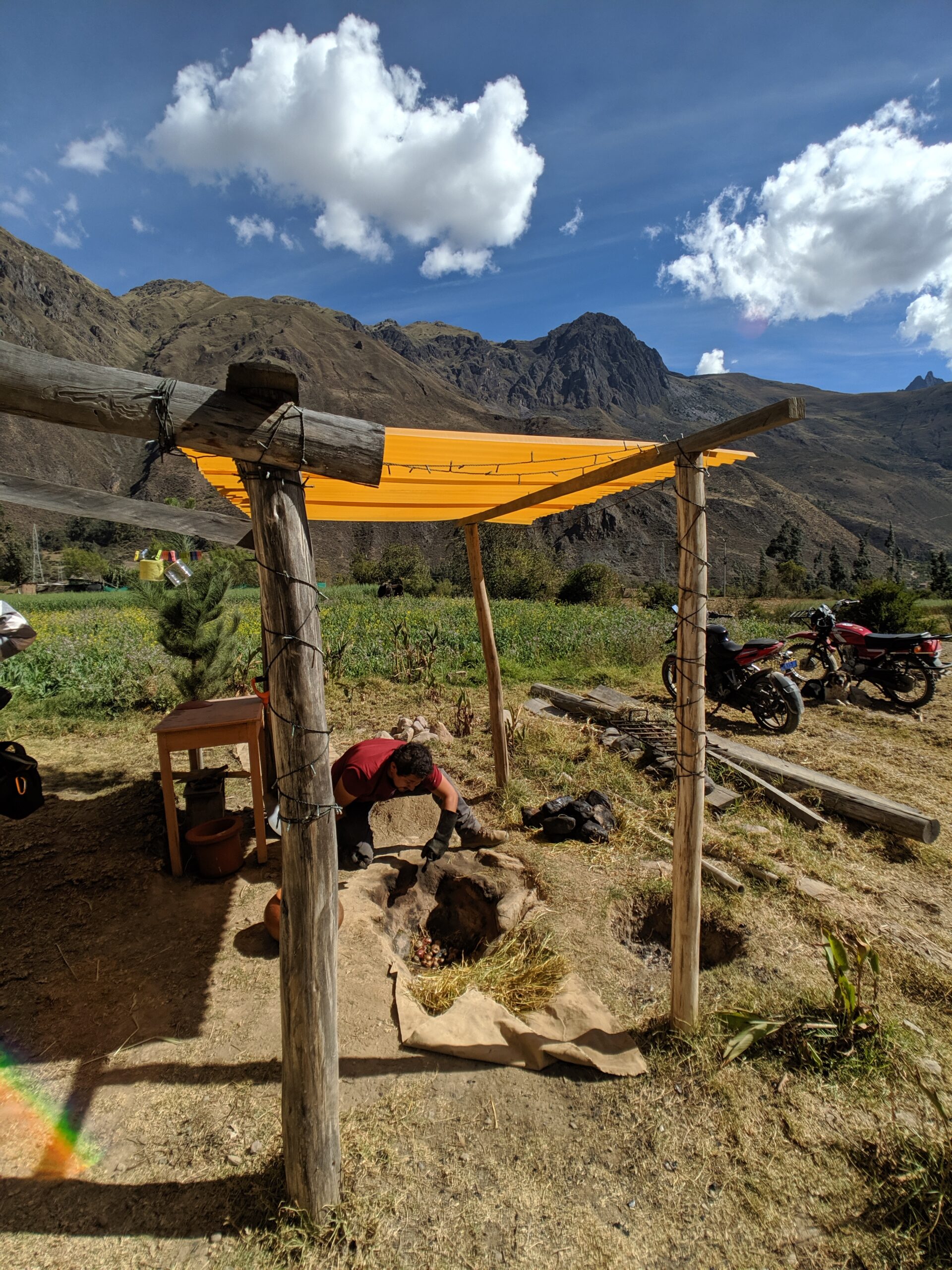 A man works to care for an underground oven under a yellow awning with a backdrop of peruvian mountains.