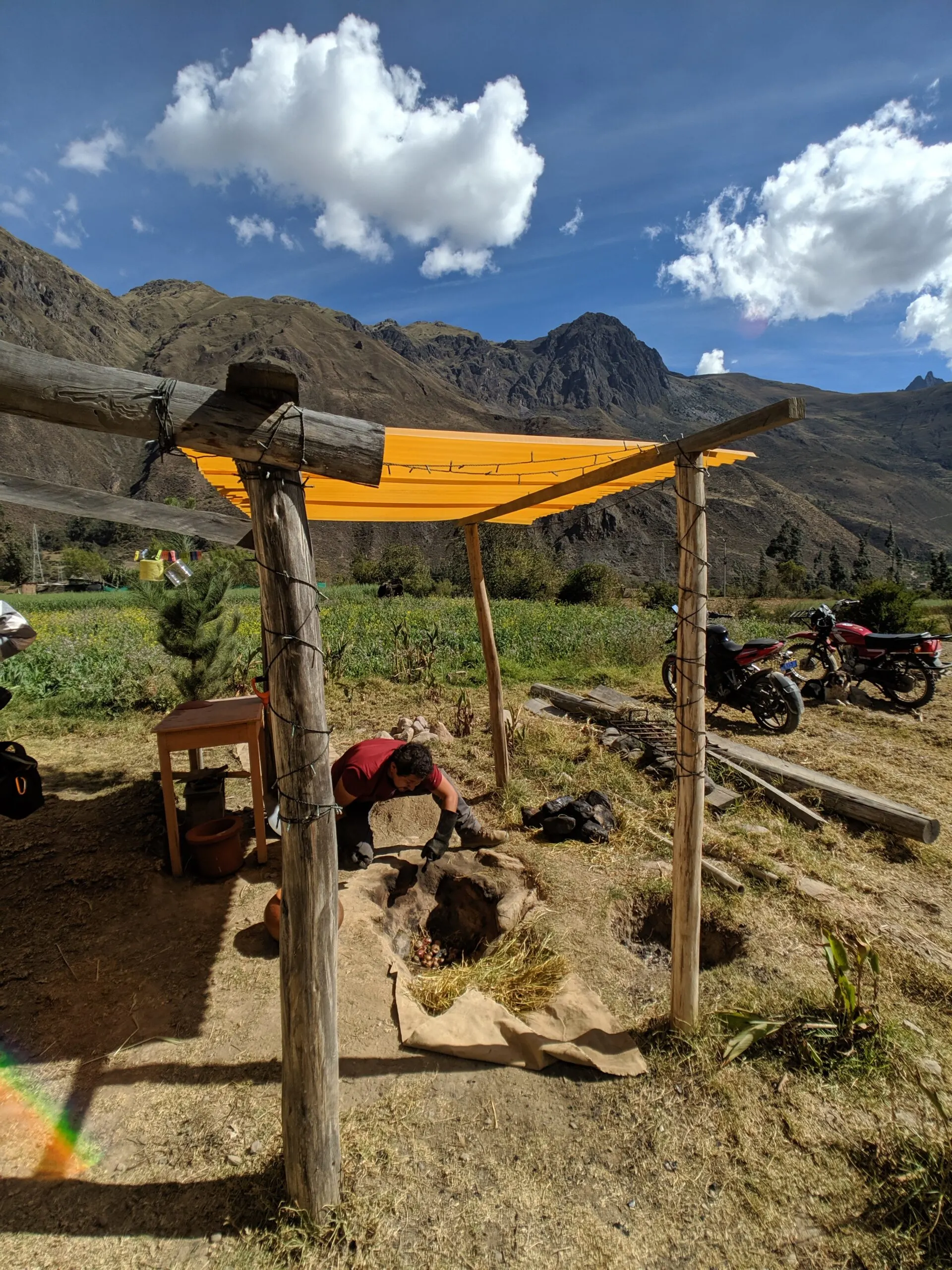 A man works to care for an underground oven under a yellow awning with a backdrop of peruvian mountains.