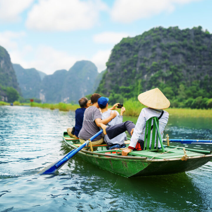 Tourists in a traditional boat in Asia snap a selfie while a tour guide paddles.