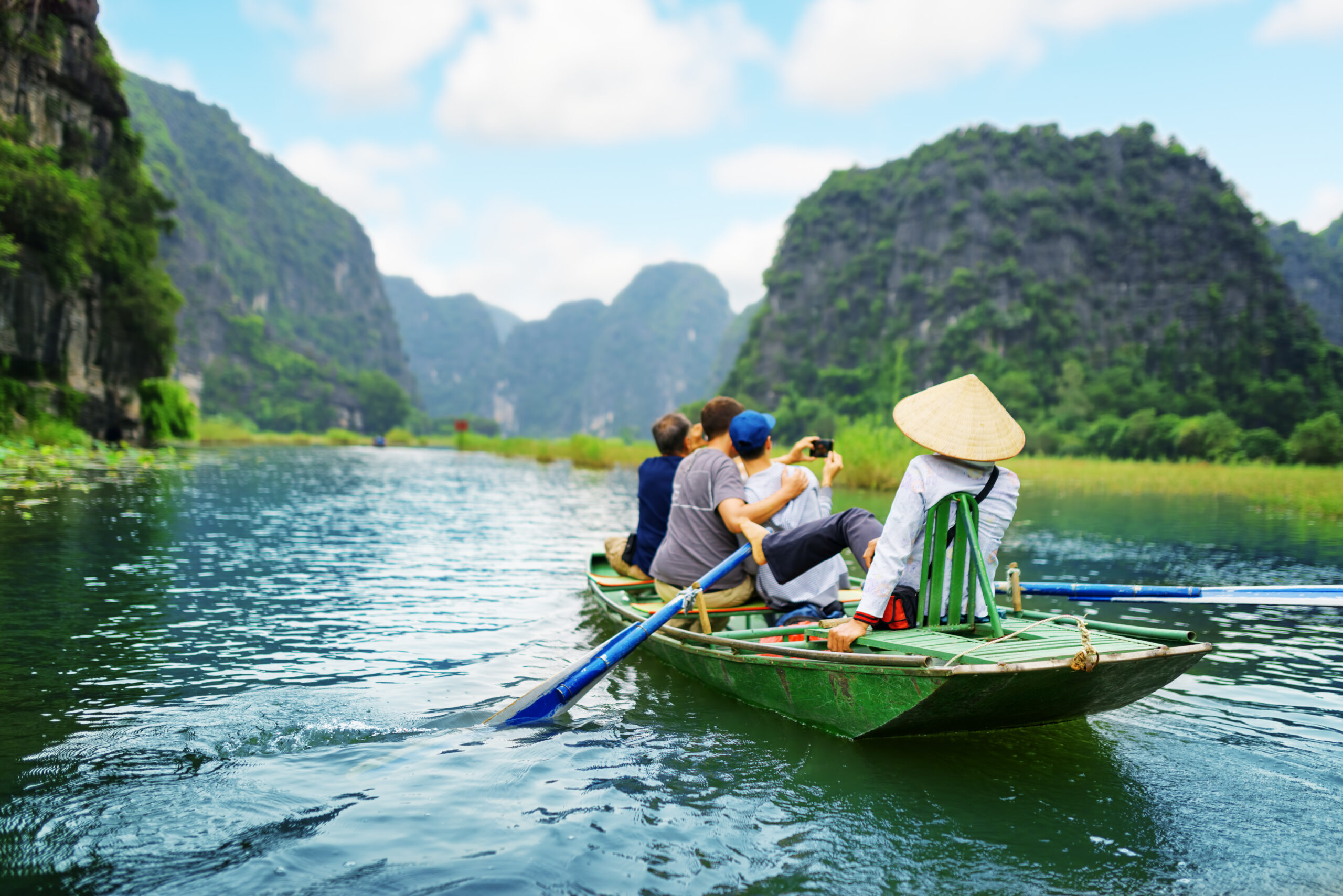 Tourists in a traditional boat in asia snap a selfie while a tour guide paddles.