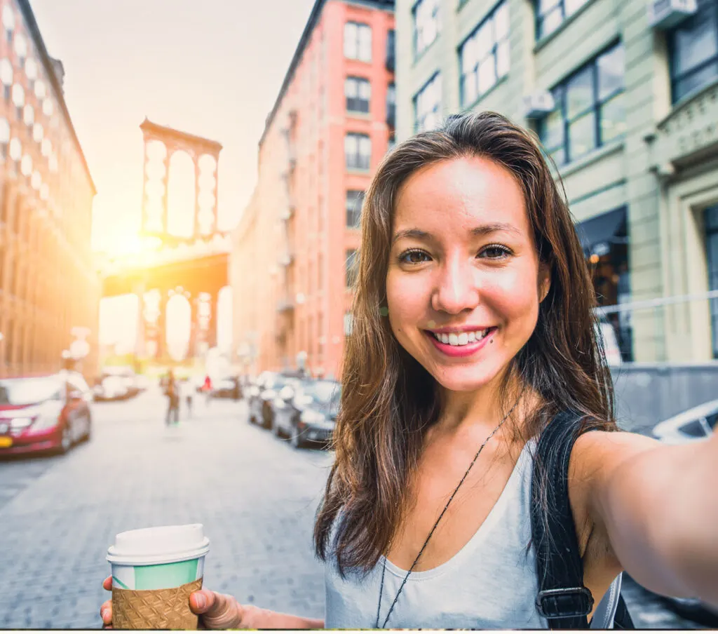 A woman stands in a street in front of the brooklyn bridge and takes a selfie photo.