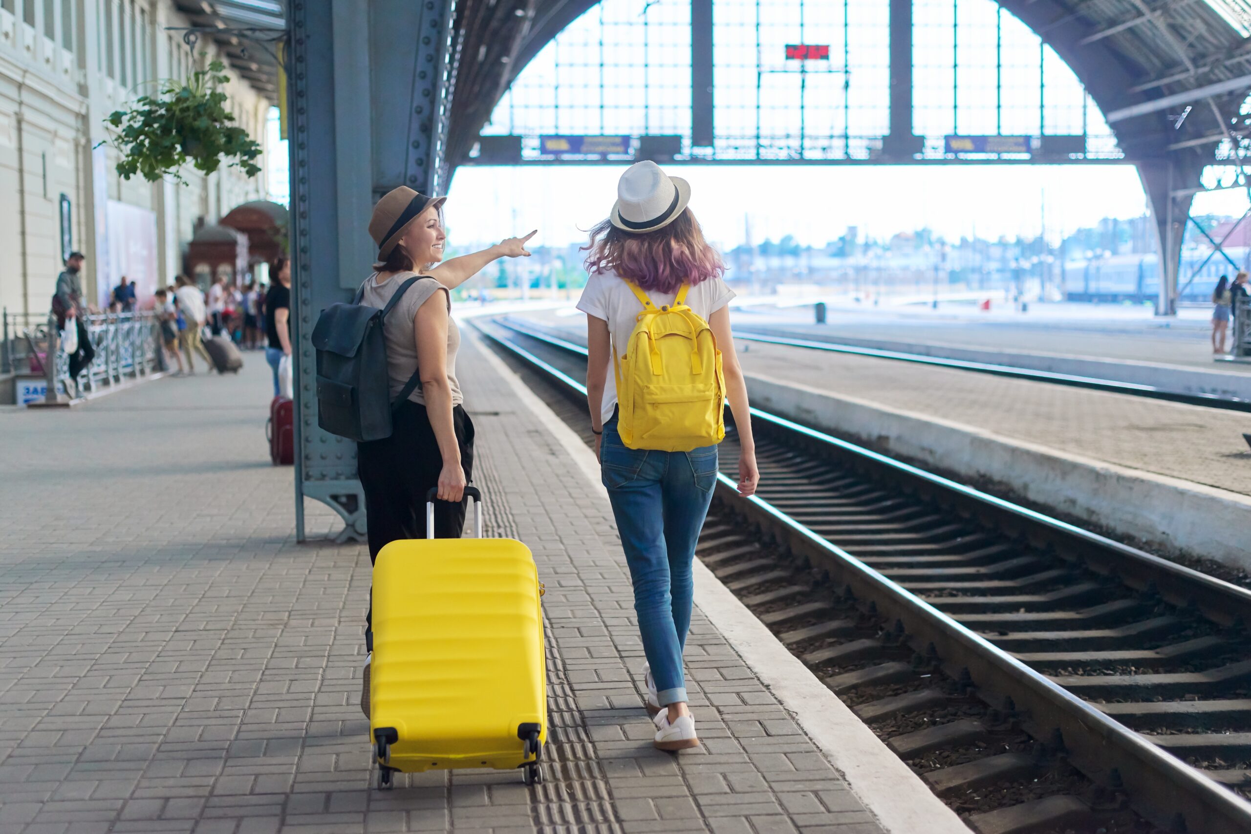 Two woman in a train station, one has a yellow suitcase and the other has a yellow backpack.