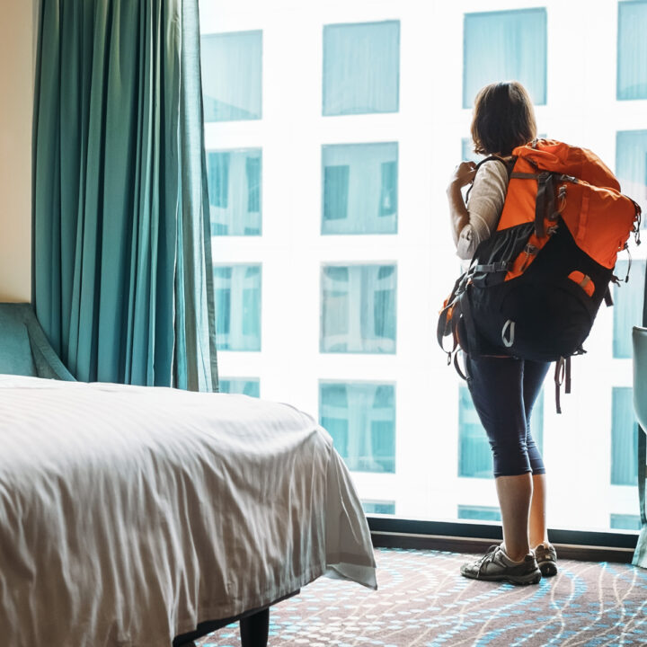 Navigating hostels without friends can be difficult, this image of a woman wearing a backpack staring out a window illustrates this concept.