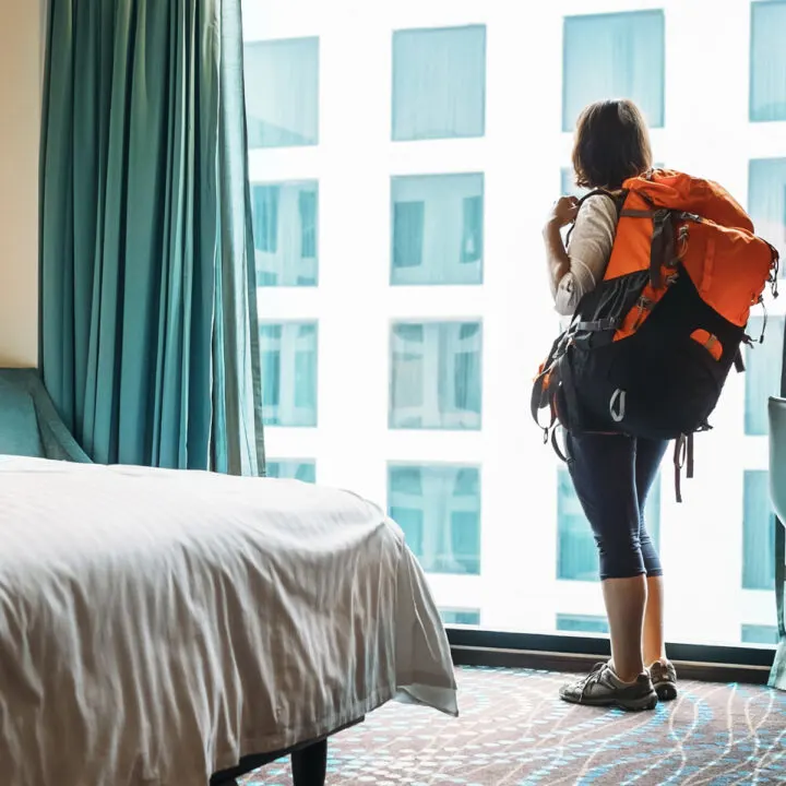 Navigating hostels without friends can be difficult, this image of a woman wearing a backpack staring out a window illustrates this concept.
