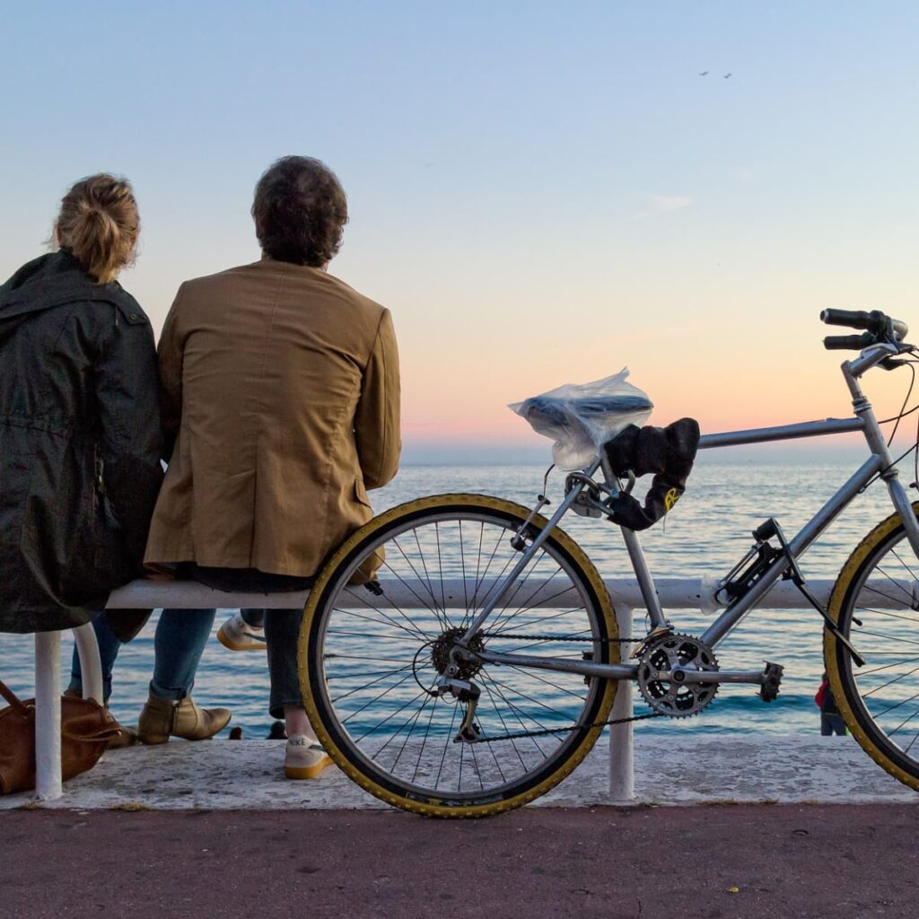 Travelers sitting on a bench near water with a bicycle nearby.