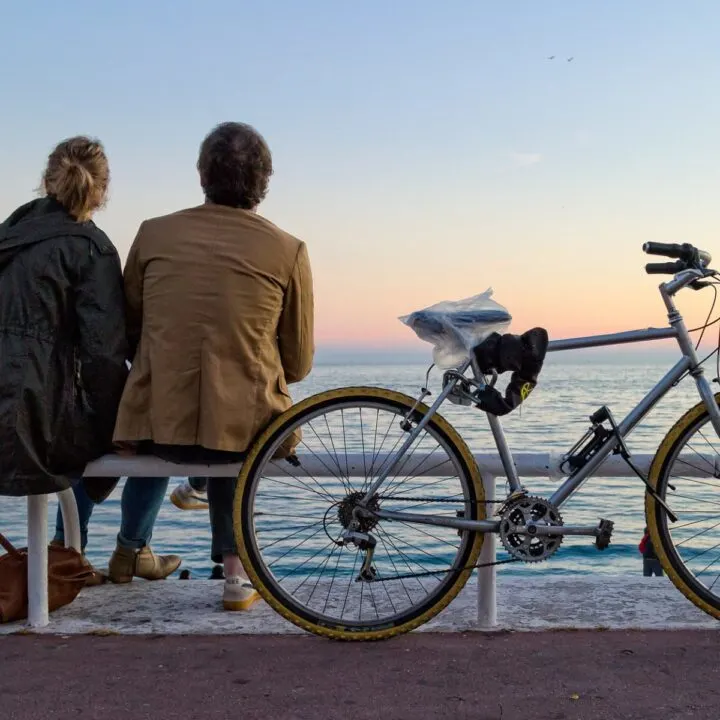 Travelers sitting on a bench near water with a bicycle nearby.