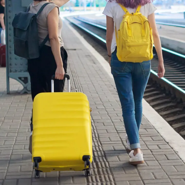 Two women walking in a train station, one woman is rolling a yellow suitcase and the other woman is carrying a yellow backpack.
