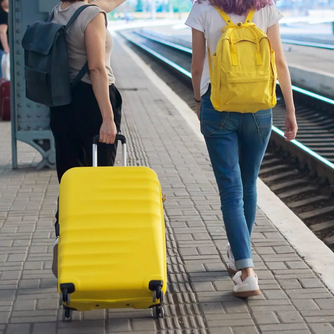 Two women walking in a train station, one woman is rolling a yellow suitcase and the other woman is carrying a yellow backpack, illustrating that even shy travelers need someone to talk with about travel.