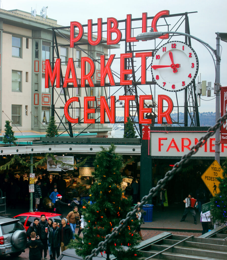 A photo of the main entrance of pike place market, showing christmas decorations, a group posing for a photo, a dancing accordion player, and the iconic neon sign.