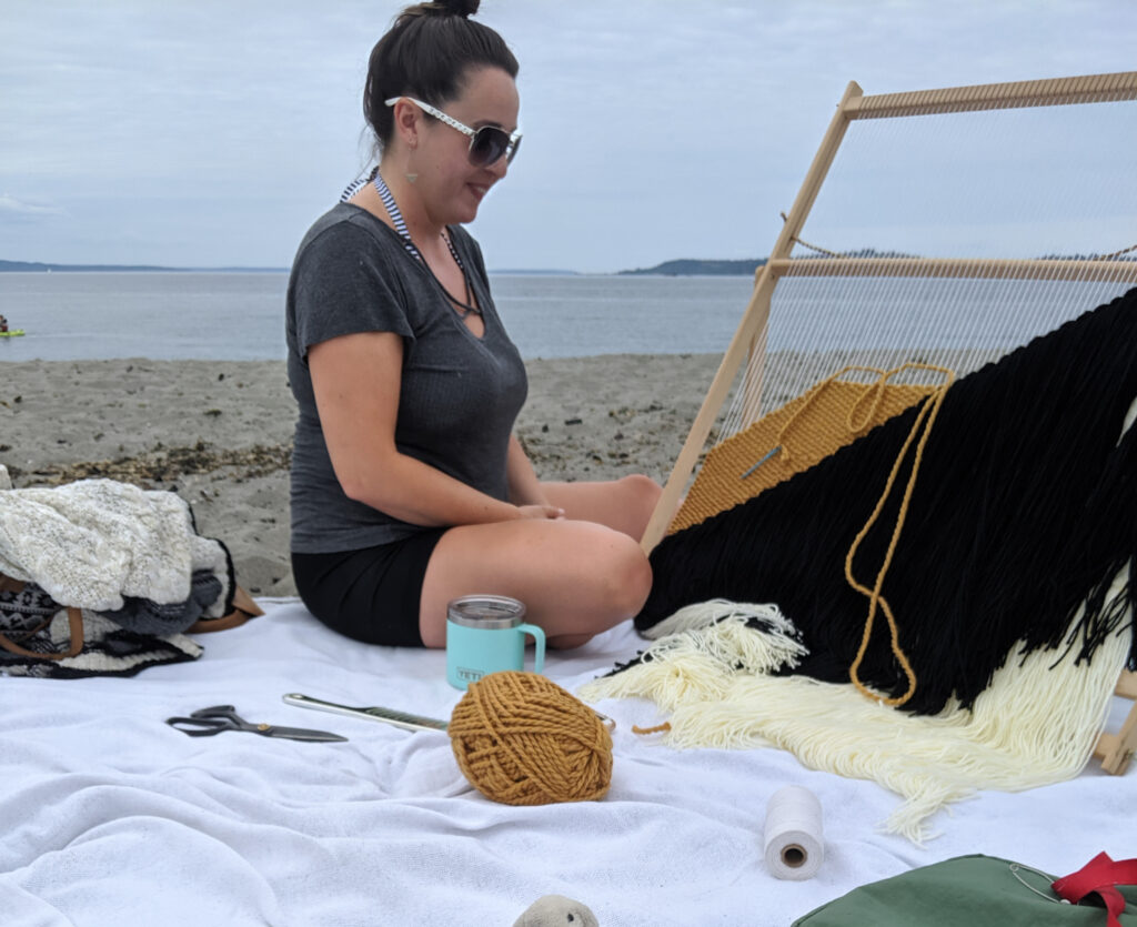 A woman sits in front of a weaving loom on a sandy beach in seattle.