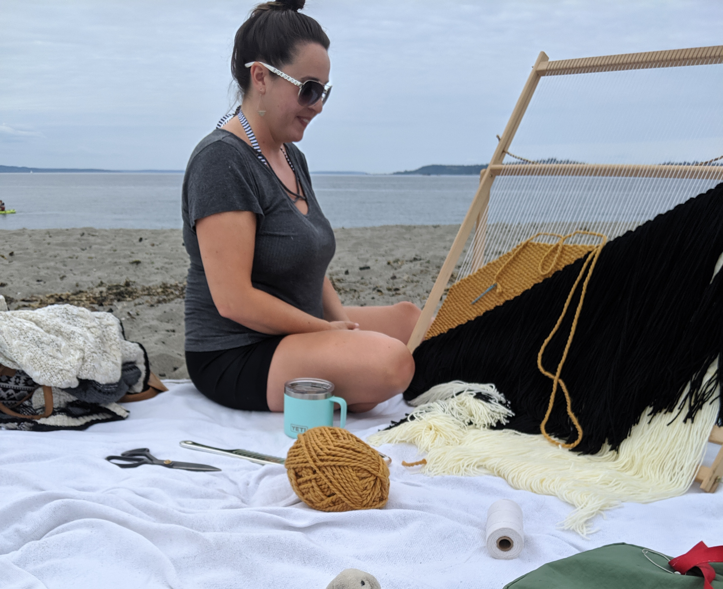 A woman sits in front of a weaving loom on a sandy beach in seattle.