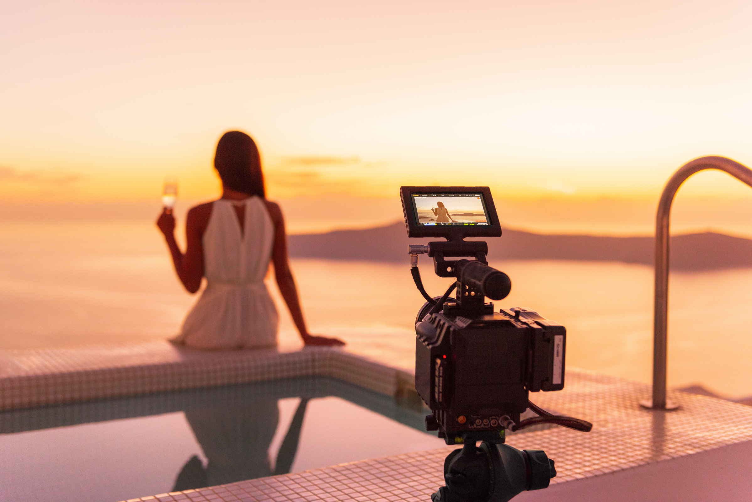 A travel influencer sits on the edge of the pool overlooking a scenic view while a camera in the forefront films her.