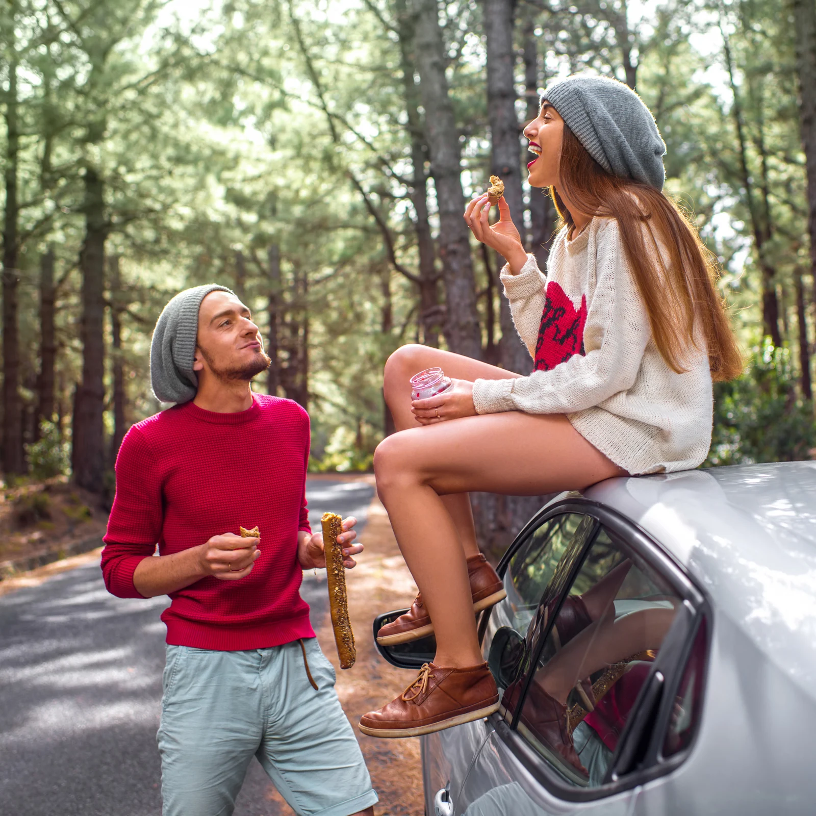 A girl sits on a car eating while a man stands nearby with the baguette.