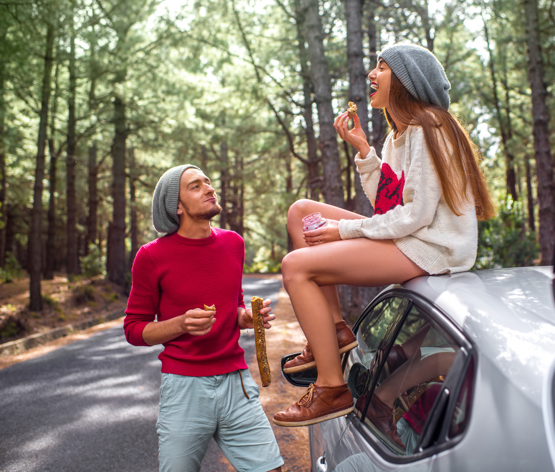 A girl sits on a car eating while a man stands nearby with the baguette.