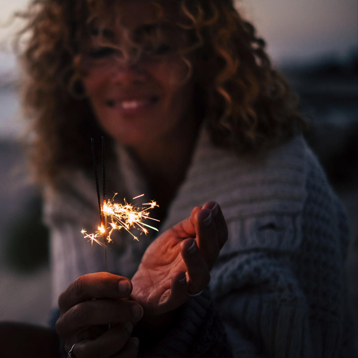 An adult woman with curly hair holding a birthday sparkler.