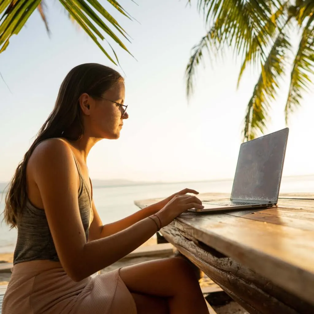 A woman types on a laptop on a beach.