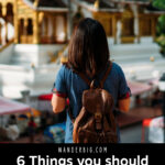 Text reading six things you should always carry while traveling overlaid over an image of a woman wearing a backpack with her back turned to the camera as she looks at a cultural building.