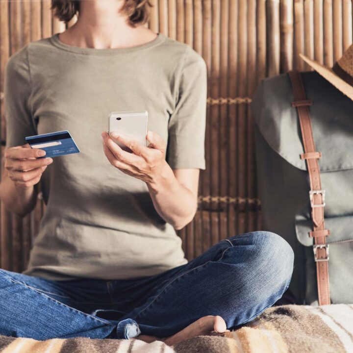 A person sits on a bed holding a phone and a credit card, next to a backpack.