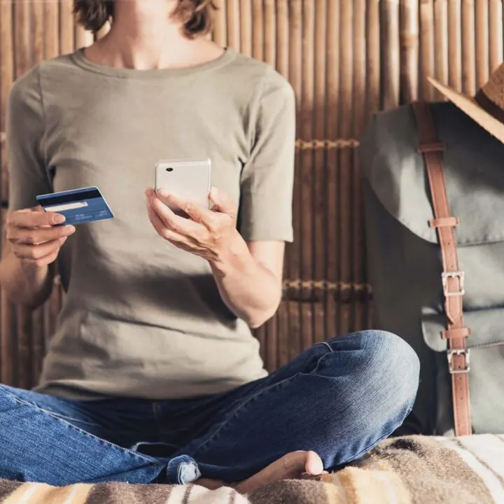 A person sits on a bed holding a phone and a credit card, next to a backpack.
