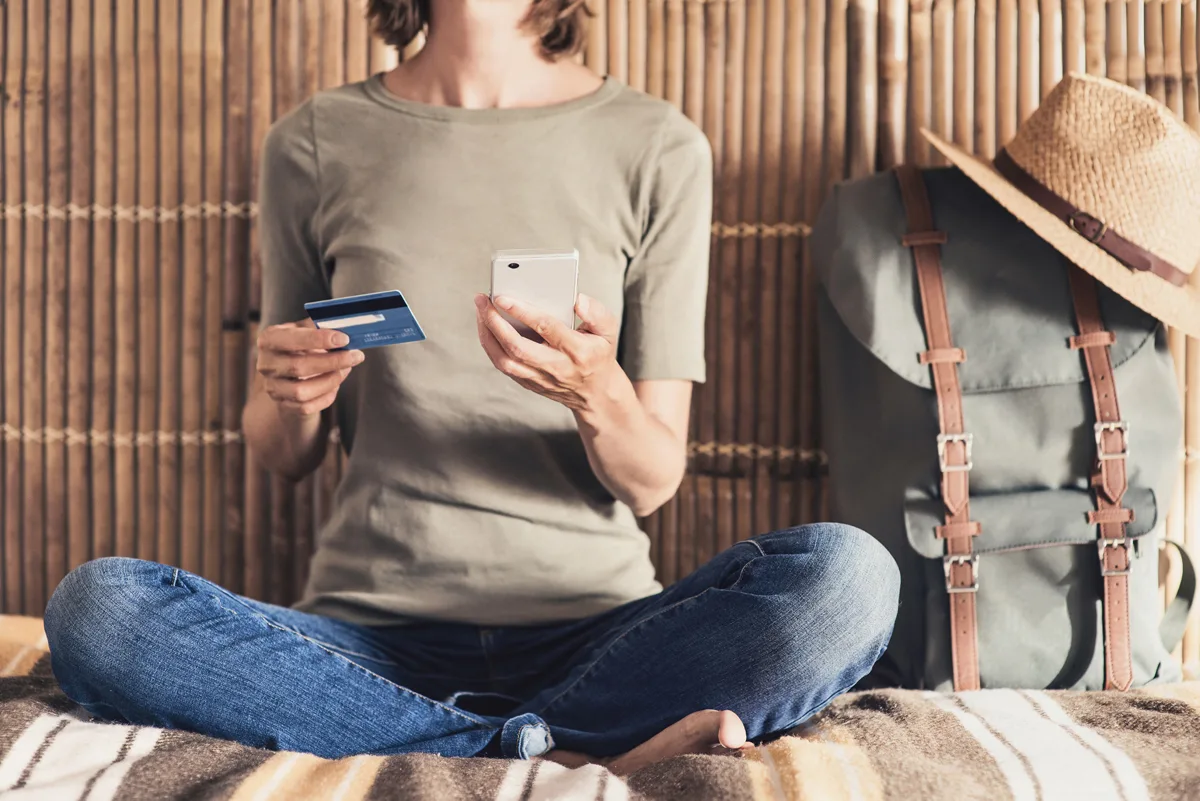 A person sits on a bed holding a phone and a credit card, next to a backpack, working on a travel budget.