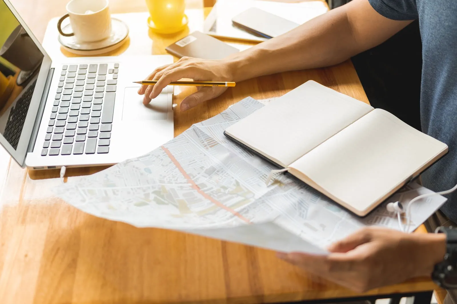 A person's forearm uses a computer while maps, notebooks, and other vacation planning supplies are spread on a table in front of them.