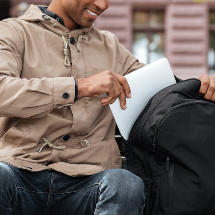 Cropped photo of young cheerful african man getting laptop from the backpack.