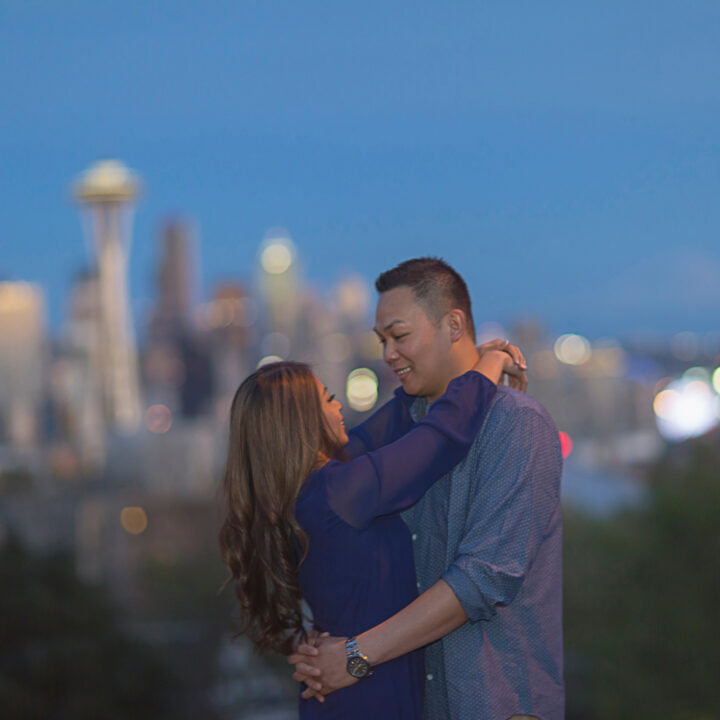 A man and a woman embrace with a blurred Seattle skyline and space needle behind them