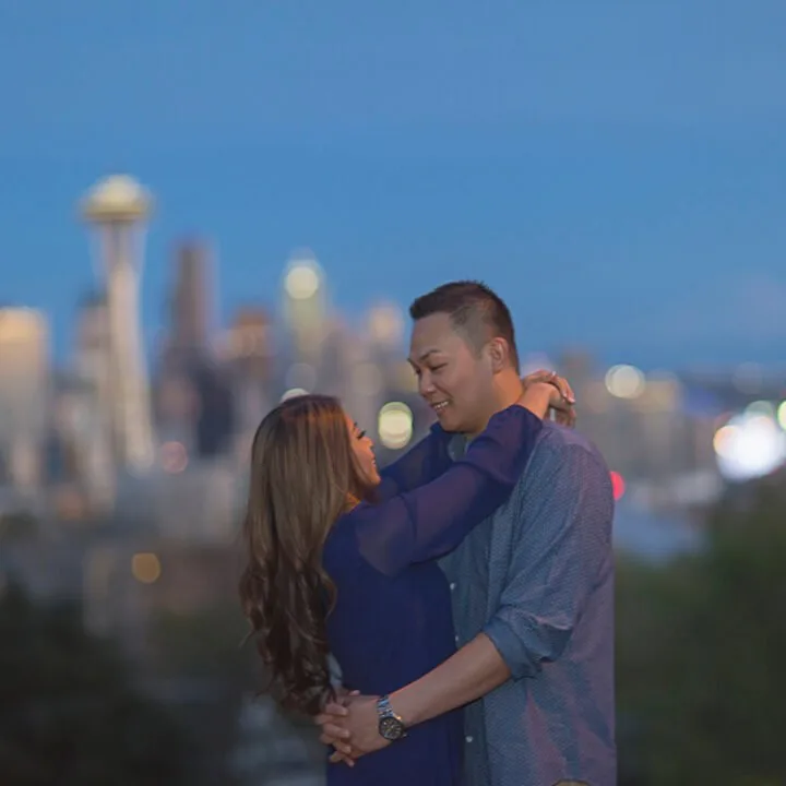 A man and a woman embrace with a blurred Seattle skyline and space needle behind them