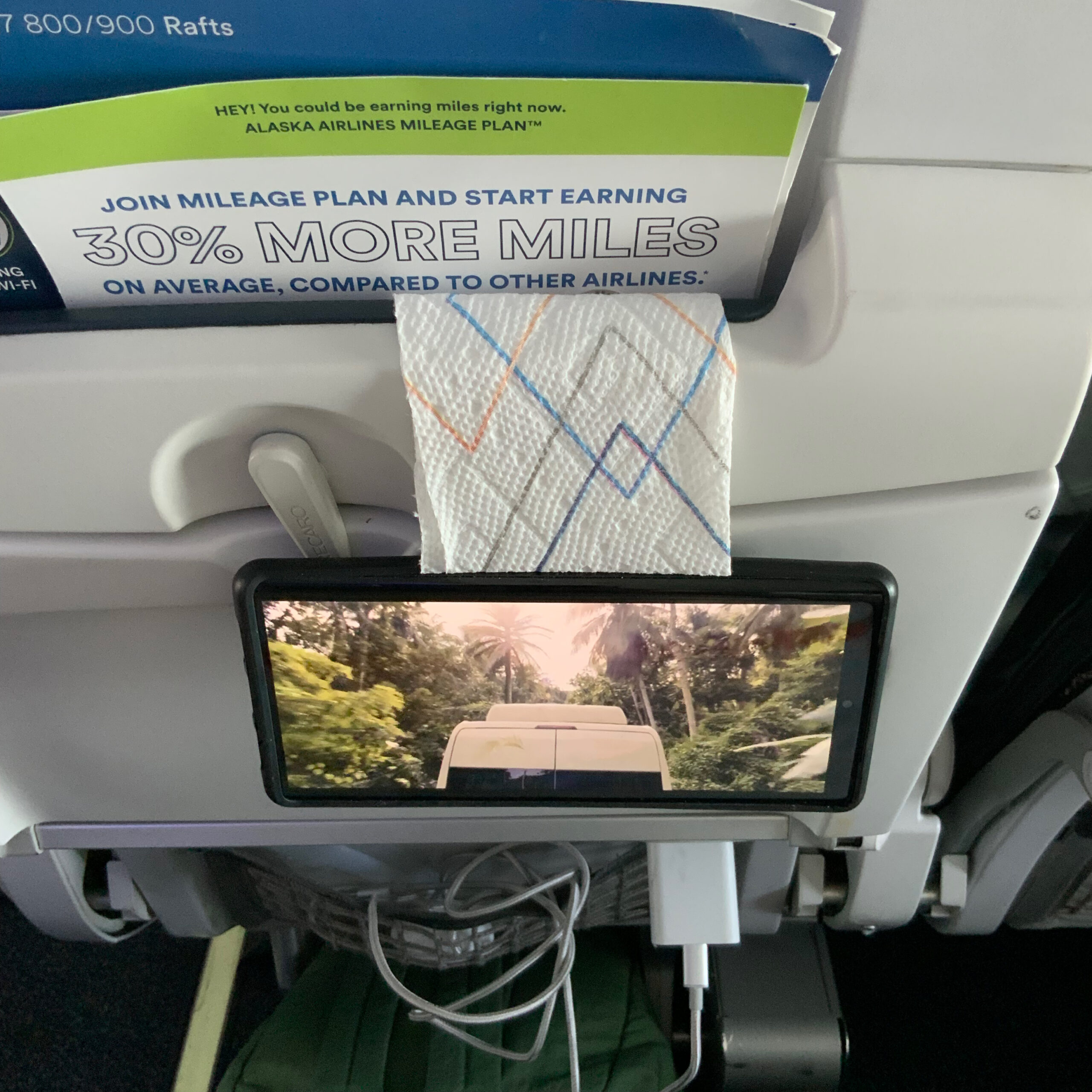 A cell phone hung from the back of an airline seat for easy viewing, set up after boarding.
