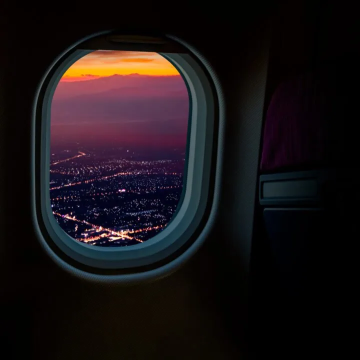 Purple mountains and city lights at sunset are shown through the window of a dark passenger compartment of an airplane.