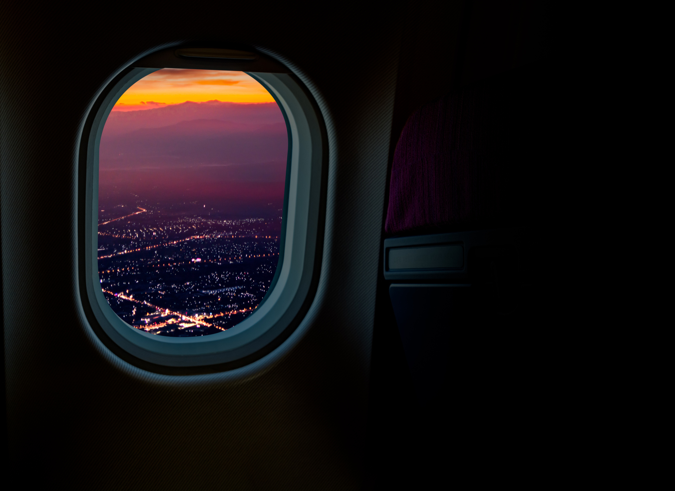 Purple mountains and city lights at sunset are shown through the window of a dark passenger compartment of an airplane.