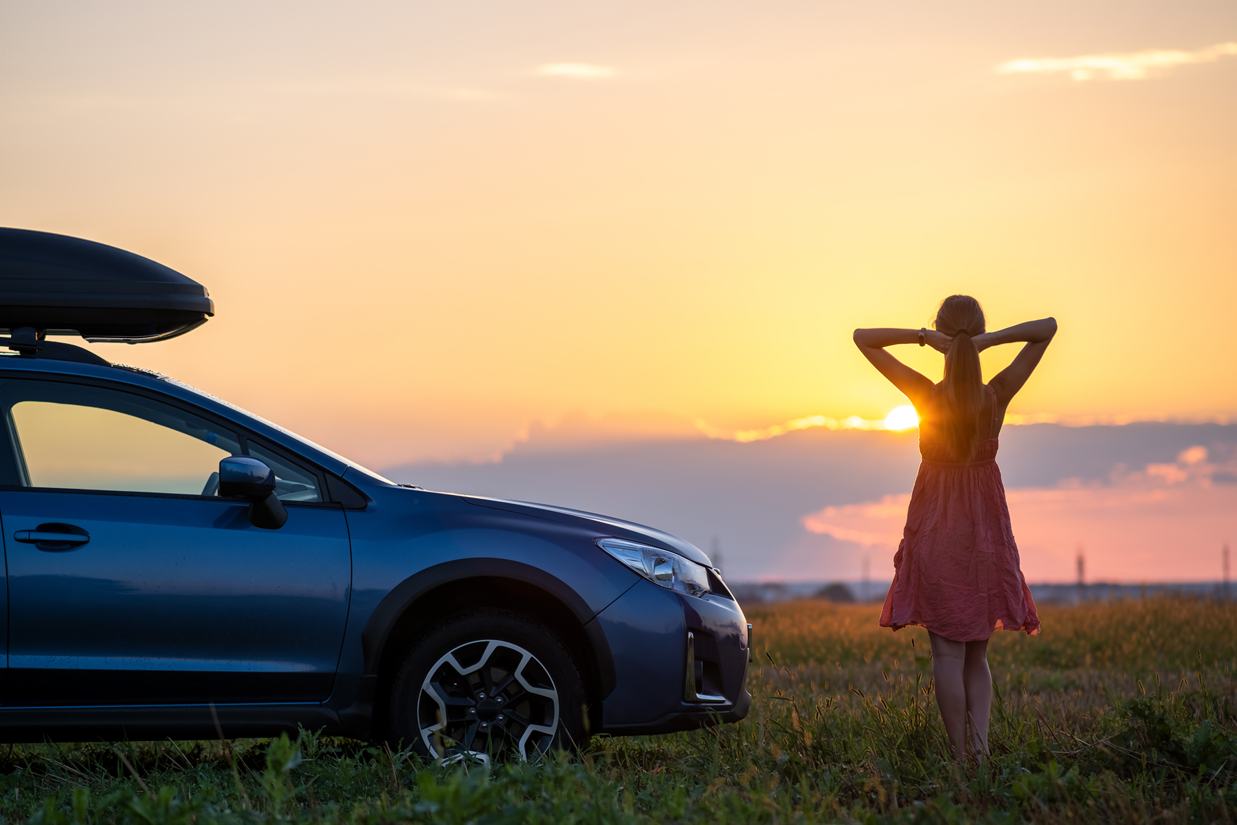 Dark silhouette of woman driver standing near her car on grassy field enjoying view of bright sunset. Young female relaxing during road trip beside suv vehicle.