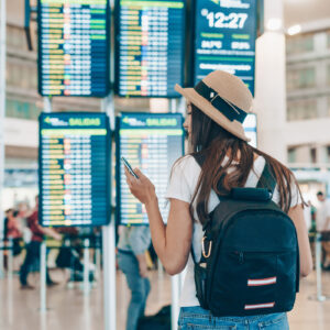 A woman with a backpack carry-on looks at a phone in front of a screen at an airport showing arrivals and departures.