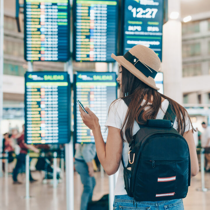 A woman with a backpack carry-on looks at a phone in front of a screen at an airport showing arrivals and departures.