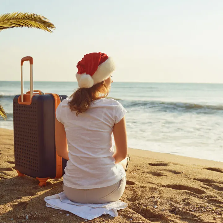 Girl wearing a christmas hat sitting on the beach next to luggage.