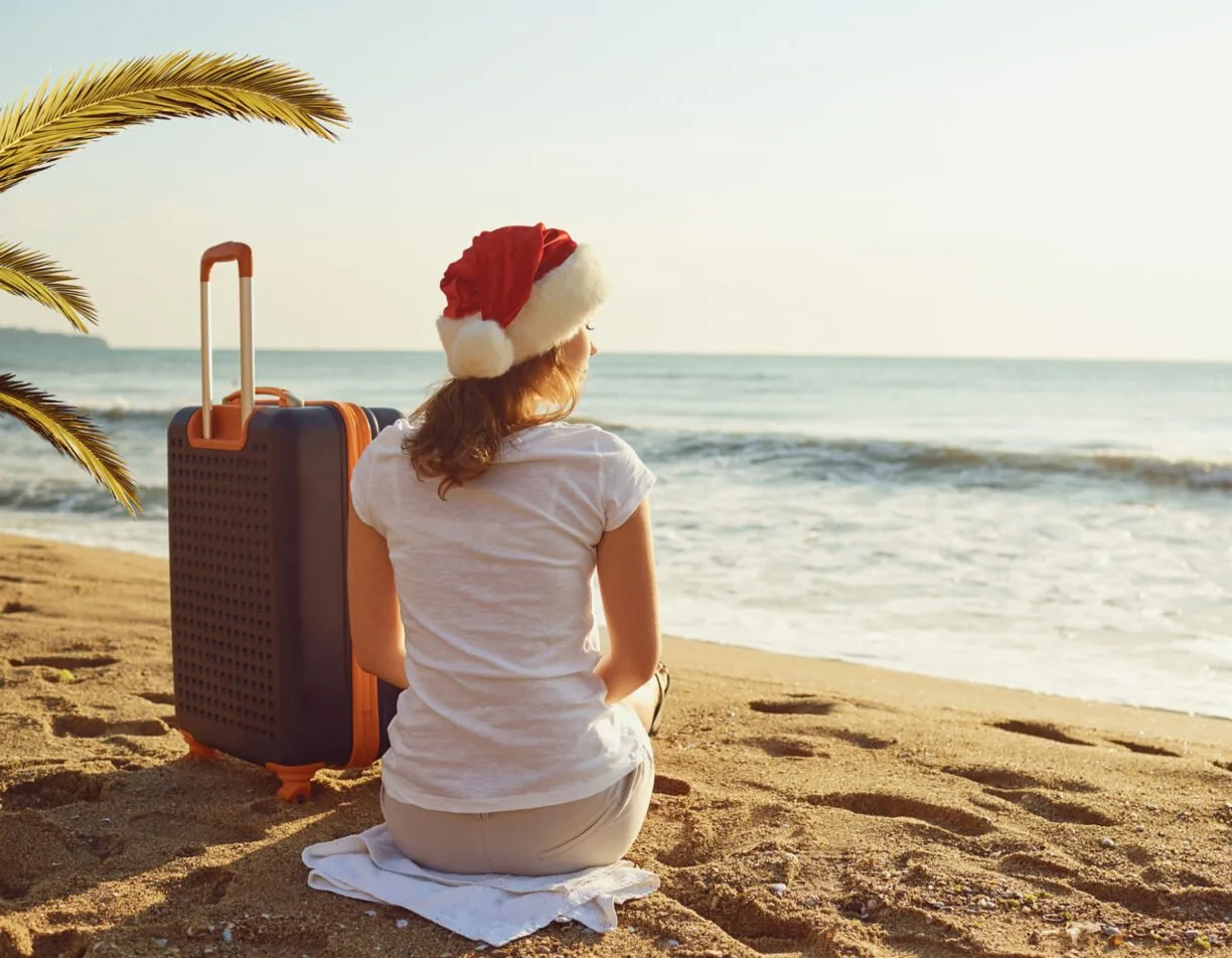 Girl wearing a christmas hat sitting on the beach next to luggage.