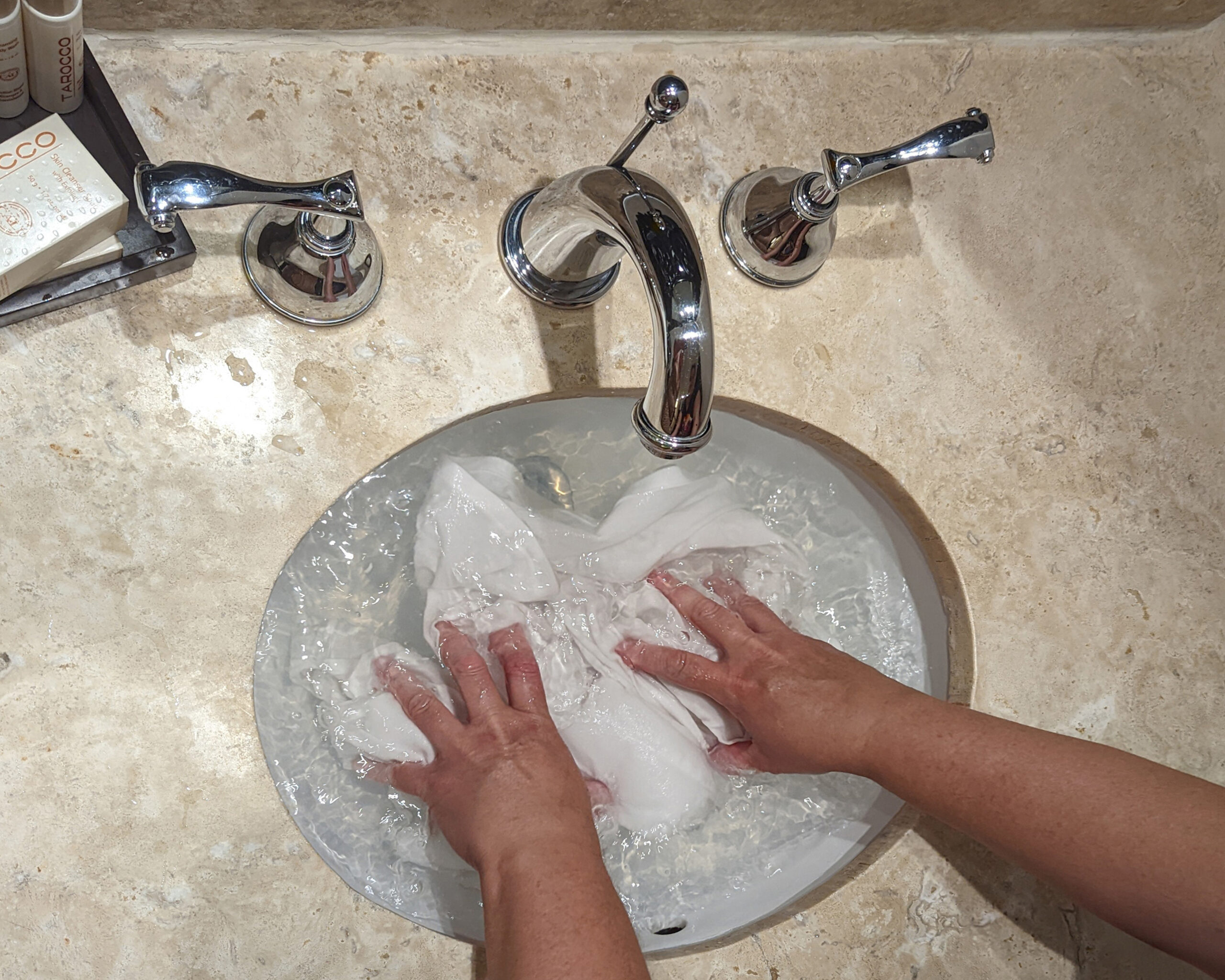 Hands washing a white shirt in a hotel sink, holding the shirt under water.
