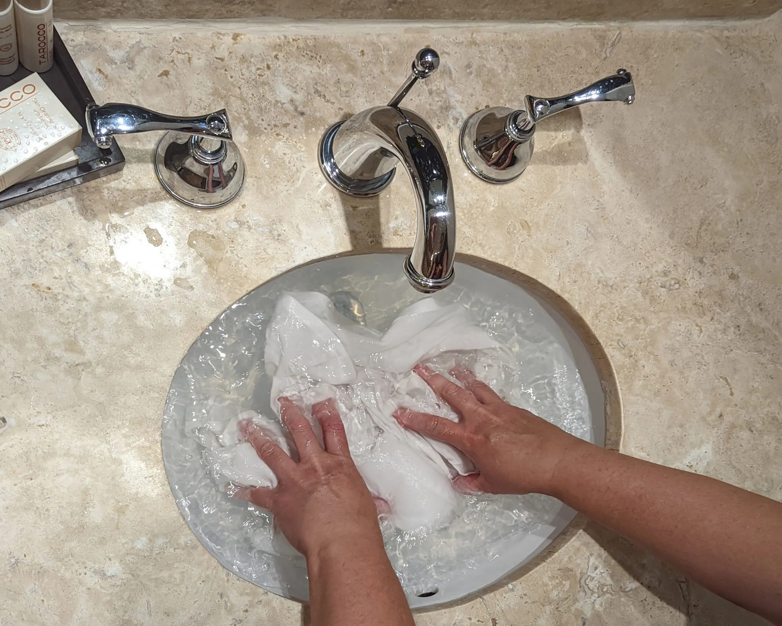 Hands washing a white shirt in a hotel sink, holding the shirt under water.