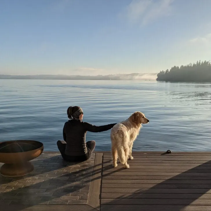 A woman and dog sit on a quiet pier in a tranquil harbor in Indianola, WA