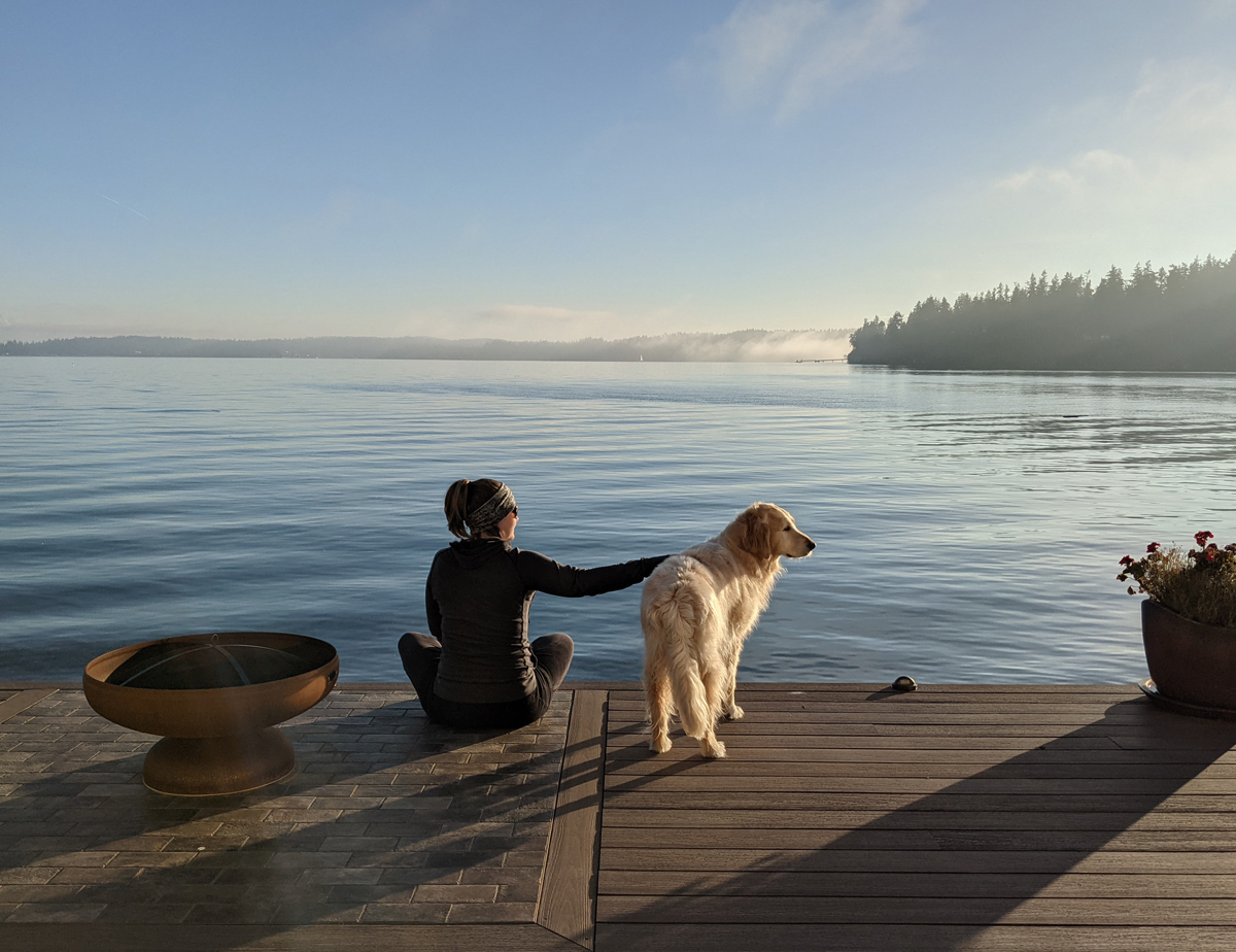 A woman and dog sit on a quiet pier in a tranquil harbor in indianola, wa