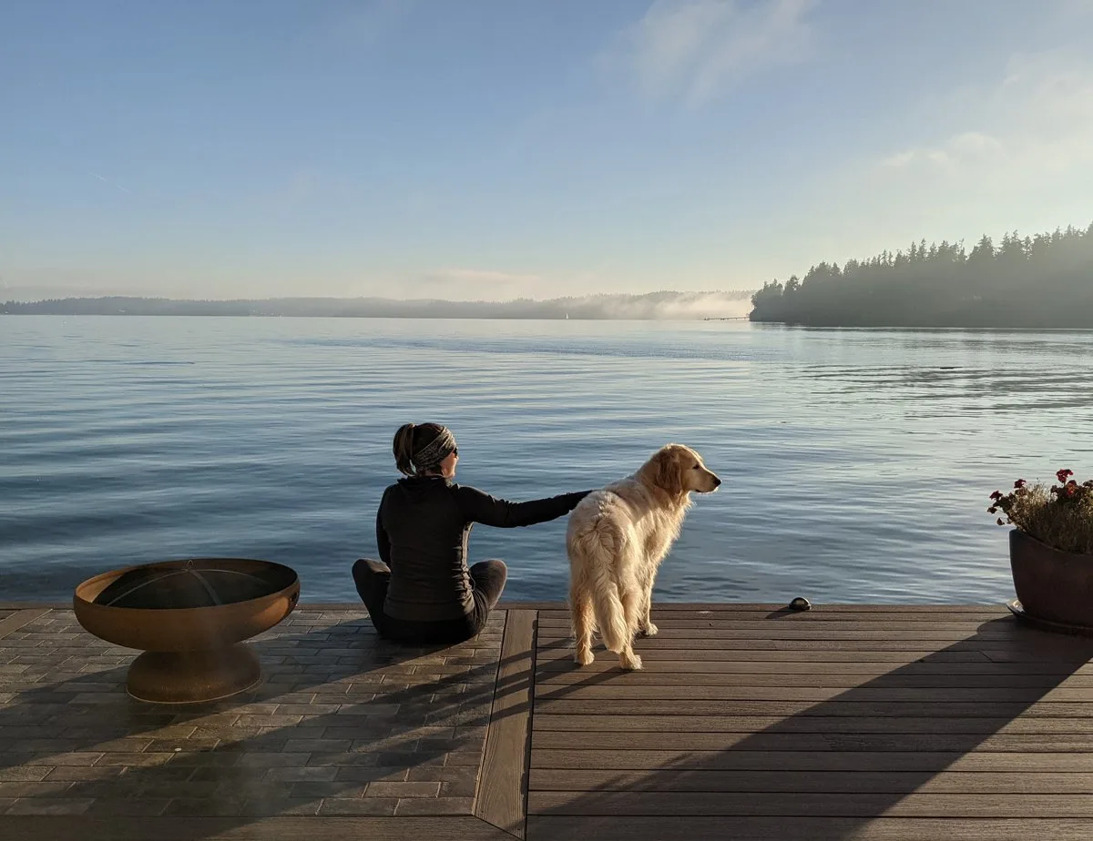 A woman and dog sit on a quiet pier in a tranquil harbor in indianola, wa