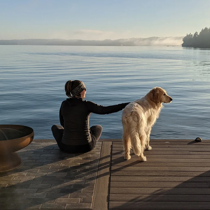 A woman and dog sit on a quiet pier in a tranquil harbor in Indianola, WA